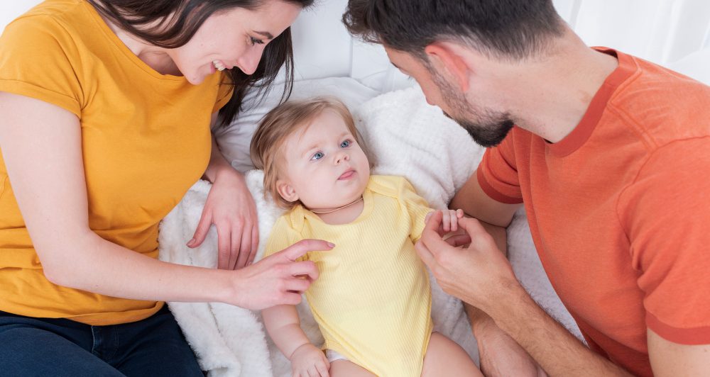 בתמונה נראים זוג הורים צעירים יושבים יחד על מיטה או משטח רך ולבן, כשהם מתקשרים בחום עם תינוקם החייכן. האם, הלובשת חולצה צהובה, מחייכת בעדינות ומלטפת את התינוק. האב, שלובש חולצה כתומה, אוחז בעדינות ביד התינוק ומביט בו בחיבה. התינוק, הלבוש בבגד גוף צהוב בהיר, שוכב נינוח בין ההורים, מביט בהם בעיניים פקוחות ובסקרנות. מסרים אפשריים של התמונה: חיזוק הקשר המשפחתי – רגעים של קרבה ואינטראקציה בין הורים לתינוקם. אהבה וביטחון רגשי – ההורים משדרים חום ותשומת לב לתינוק. תקשורת הורית חיובית – ההורים מתקשרים עם התינוק דרך מגע, מבטים ודיבור. רוגע ושקט נפשי – הסביבה הבהירה והרגועה משדרת שלווה וביטחון